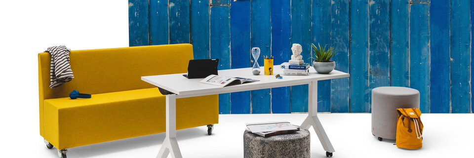 Gispen JUNA Single low trainbench upholstered in yellow with castors and a white Dukdalf folding table with 2 grey Asido poufs in front of blue scrapwood backpanel front left view