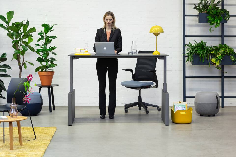 Royal Ahrend Balance workstation in grey with charcoal top in stand position and 2020 office chair with female model standing front view