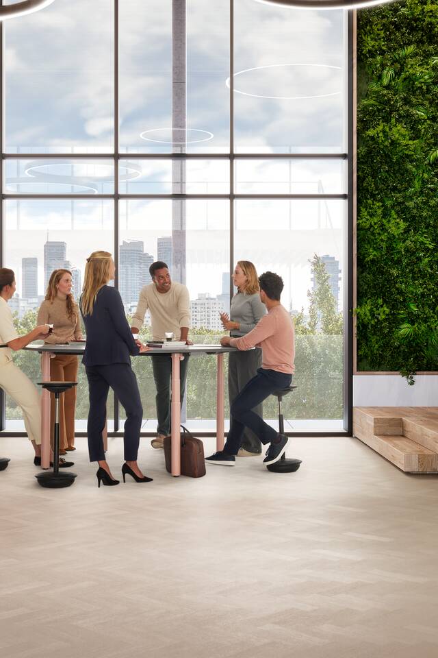 Ahrend Modea sit stand conference table in pink with grey round tabletop in stand height with sitting and standing models in a vitalising workspace interior view