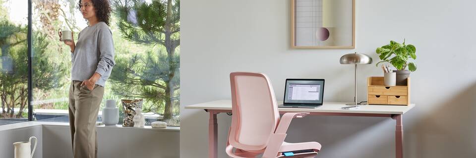 Ahrend Remode office chair in pink with balance solo workstation and model standing holding cup in a home setting right rear view