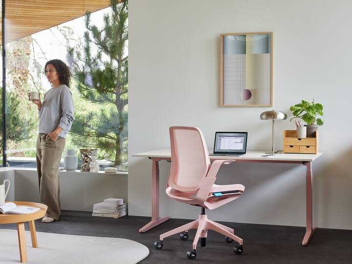 Ahrend Remode office chair in pink with balance solo workstation and model standing holding cup in a home setting right rear view