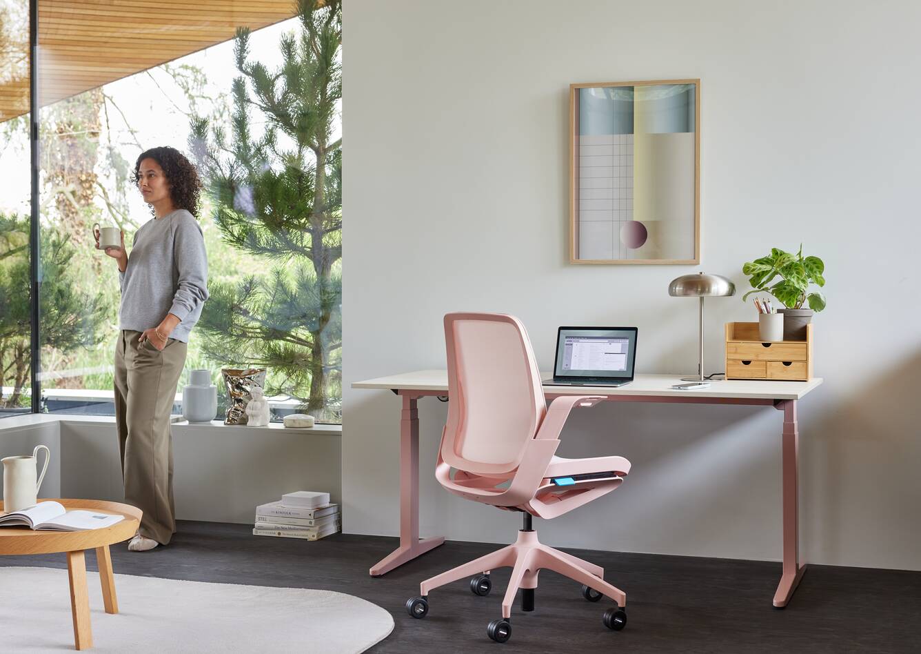 Ahrend Remode office chair in pink with balance solo workstation and model standing holding cup in a home setting right rear view