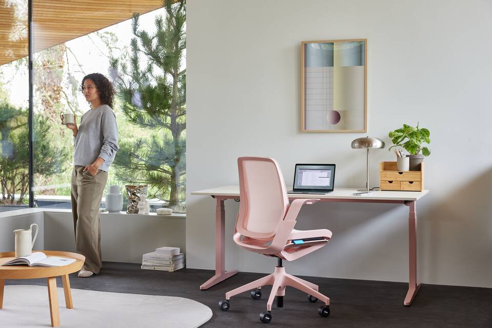 Ahrend Remode office chair in pink with balance solo workstation and model standing holding cup in a home setting right rear view