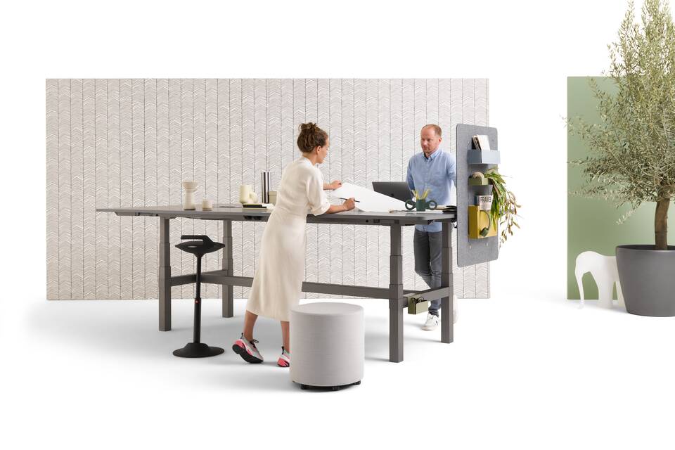 Gispen TMNL Agile table in dark grey with grey tabletop in standing position and backpanels with 2 models with grey Asido pouf styled front right view