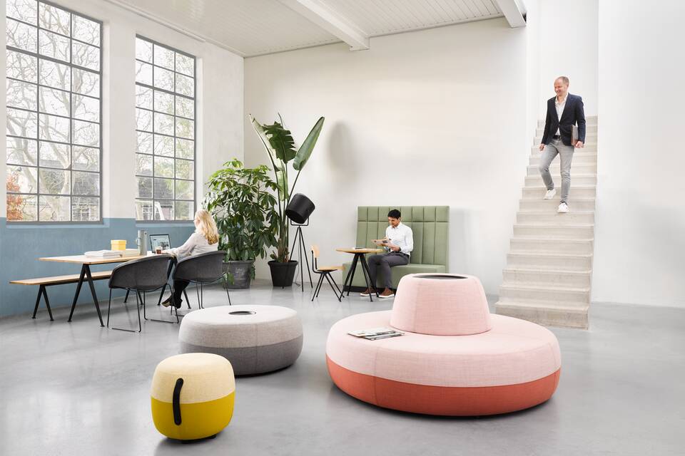 Ahrend Recharge poufs upholstered in pink yellow and grey with Pyramid and Well armchairs with models in a Hybrid working community setting