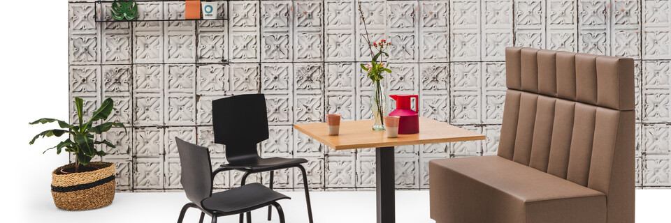 Gispen JUNA single high trainbench upholstered in brown with black KOLM column table and 2 black More or Less chairs in front of grey patern backpanel front right view